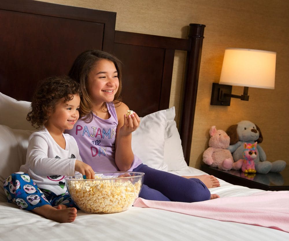 Big sister and little sister watching Tv and eating popcorn in the Hyatt Bellevue hotel room.