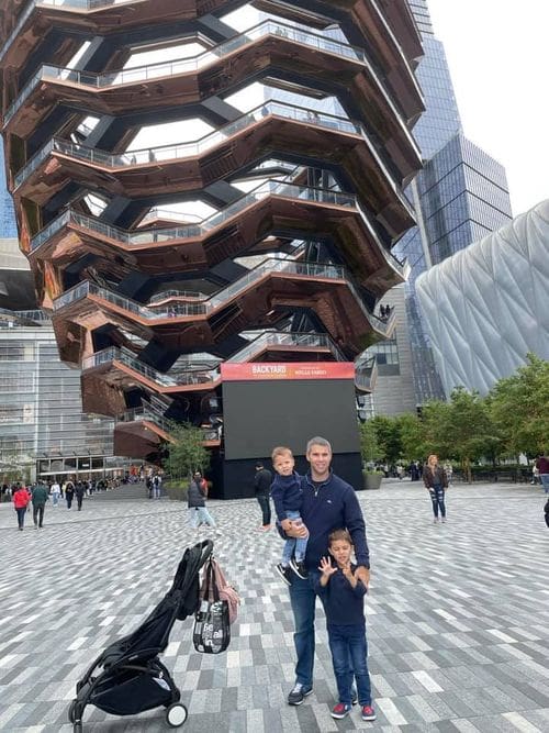 A dad and his two kids stand together with the Vessel in Hudson Yards behind them.