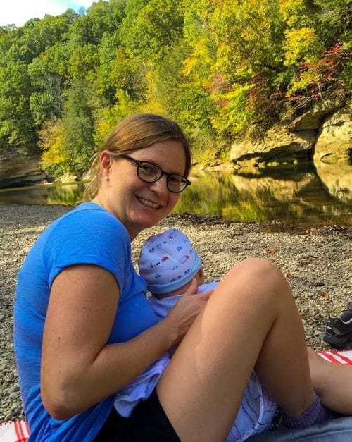 A mom holds her young son on a rocky beach, with fall colors on the far shore, in a state park in Parke County, Indiana.