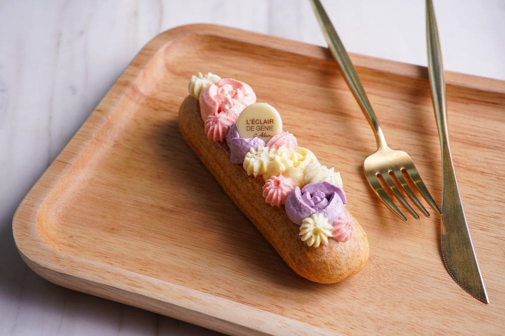 A decorated eclair rests on a wooden board, featuring white, purple, and pink frosting on top. A golden-hued knife and fork rest nearby.