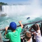 Three kids stand at a railing looking over it into Niagara Falls, where a boat is on the water headed toward the falls.