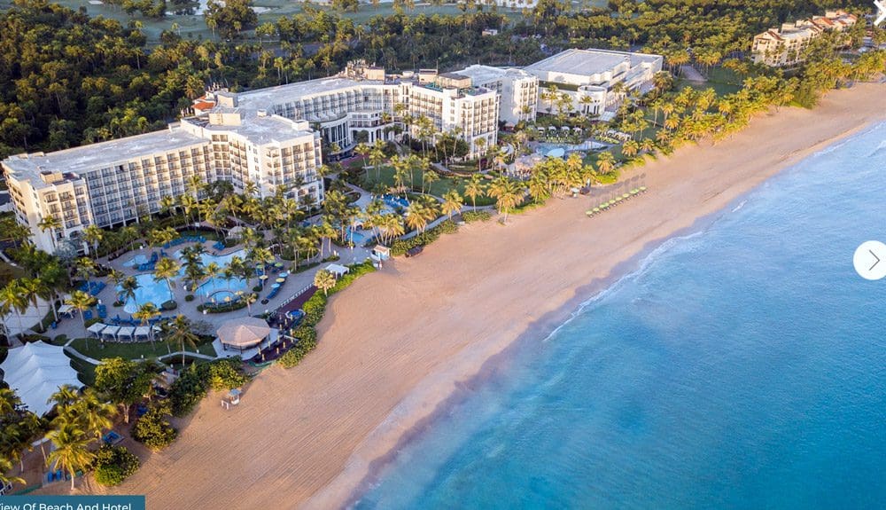 Aerial view of the beachfront where the Wyndham Grand Rio Mar Puerto Rico Golf & Beach Resort is located. Shows the hotel pools, palm trees, and a beautiful blue ocean. 