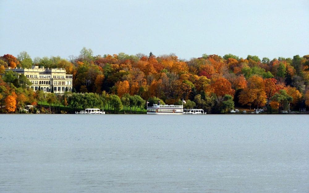 The shoreline of Lake Geneva, featuring a mansion on shore, and a boat in the water, along with brilliant fall foliage in hues of orange, red, and green.