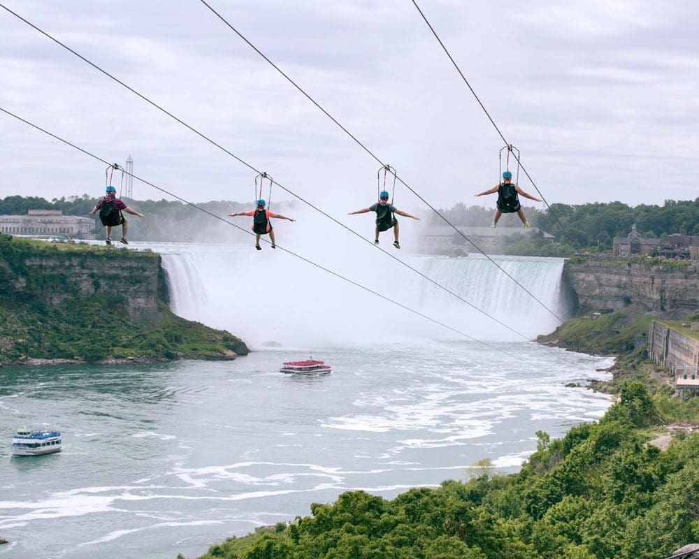 Four people zip-line at WildPlay Niagara Falls, with a view of Niagara Falls behind them.