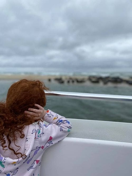 A young girl with red hair looks over a boat rail toward the seals lounging on Monomoy Island.