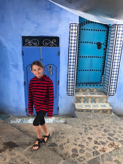 A young girl stands in front of a blue door in Chefchaouen, Morocco.
