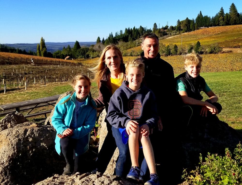 A family of five sits together on large boulders with a large winery behind them.