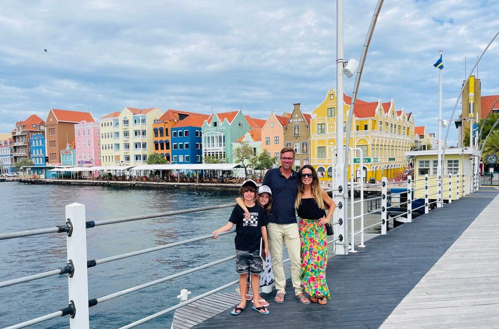 A family of four poses together along a boardwalk with colorful buildings from the city of Punda in the distance.