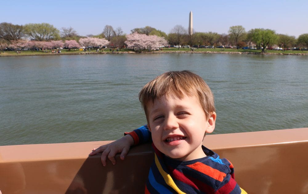 A young boy enjoys a boat ride along the river in Washington DC, with views of cherry blossoms in the distance.
