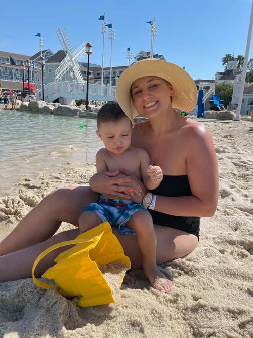 A mom holds her son, both wearing swim suits, on the beach near the Disney Yacht Club.
