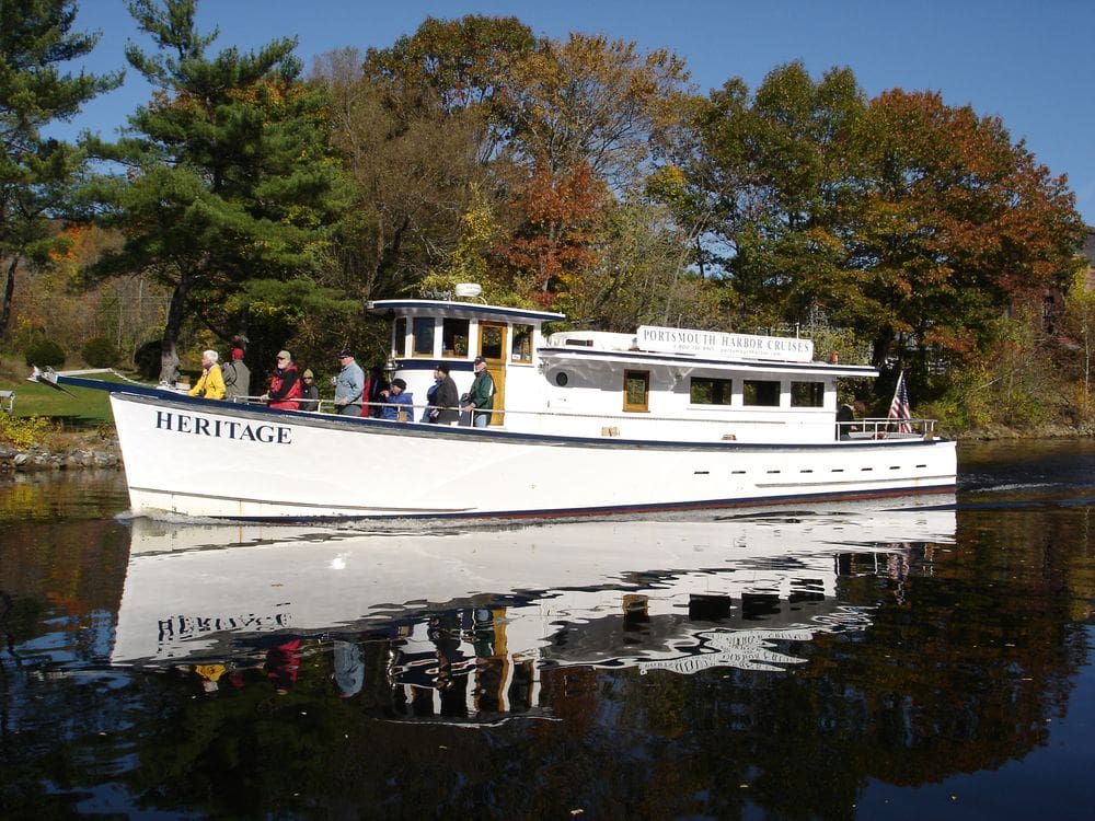 A white boat with the name "Heritage" floats down the river on a beautiful autumn day with several passangers and fall foliage along the shore.