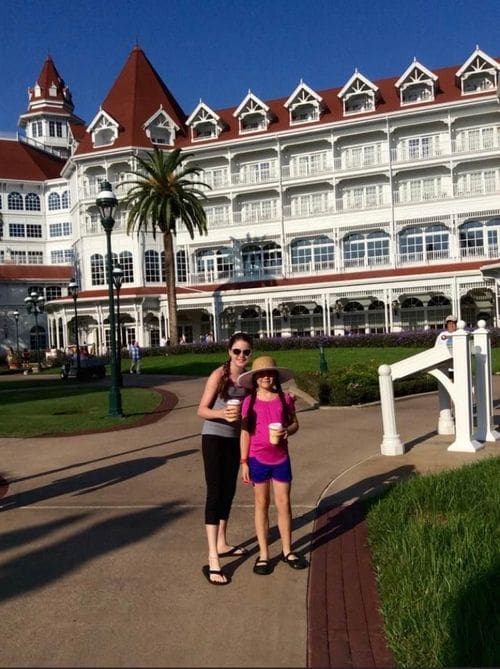 A mom and her daughter, both holding warm beverages, stand together outside the Grand Floridian, with its iconic red roof and white exterior and balconies.