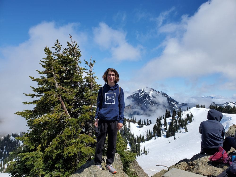 A teenage boy stands on a rock with tall pines and a snowy mountain behind him at Hurricane Ridge, one of the best hikes near Seattle for families.