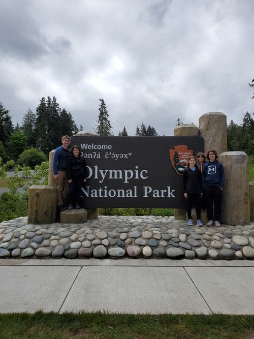 A family of five stands near a national park sign reading "Olympic National Park".
