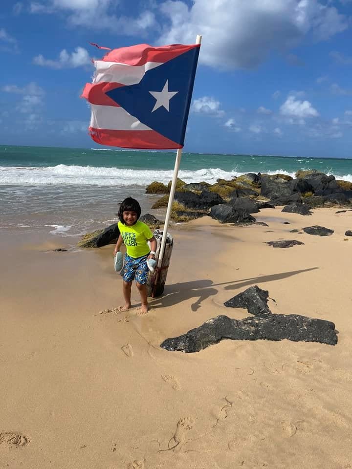 A young boy stands on a beach with a Puerto Rican flag waving in the wind near him.
