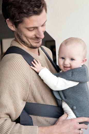 Close up of a dad smiling at his child, while holding the child in a dark gray front carrier by BABYBJÖRN Baby Carrier Mini.