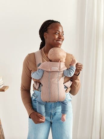 An African-American mom holds her infant in a blush pink front carrier by BABYBJÖRN Baby Carrier Mini, one of the best baby carriers for travel.