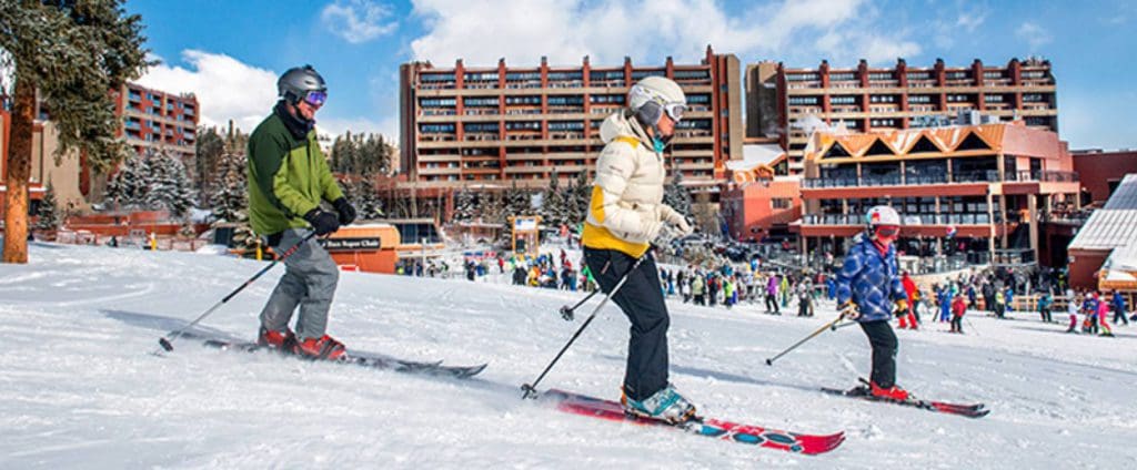 A family of three skis down a slope with a view of Beaver Run Resort & Conference Center behind them.