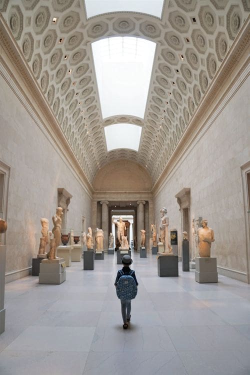 A young boy stands in the middle of a long hallway, featuring a variety of sculptures, at the Met.
