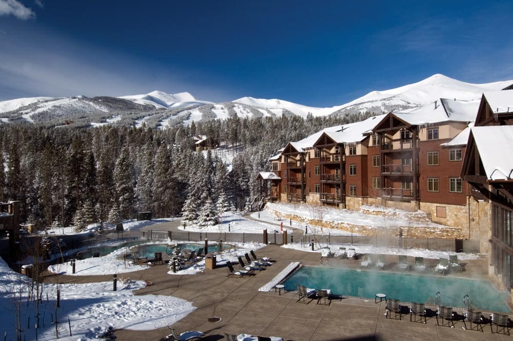 A view of the resort buildings and grounds at Grand Timber Lodge, one of the best places to stay in Breckenridge with kids this winter, featuring two pool areas, and a snow-covered mountain in the distance.