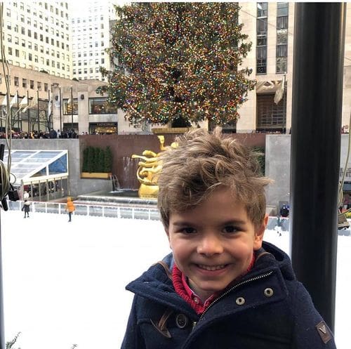 A young boy smiles with an NYC skating rink behind him.