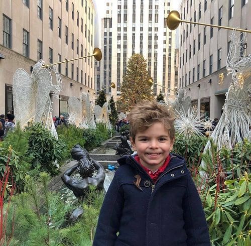 A young boy stands smiling with the Rockefeller Plaza behind him, decked out in Christmas decor.