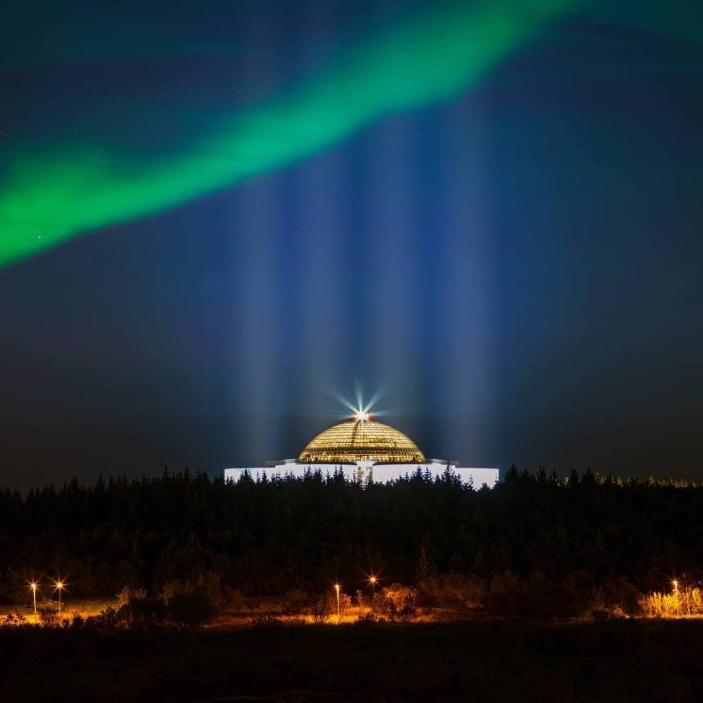 The Perlan Museum can be seen lit up in the distance, with green northern lights overhead.