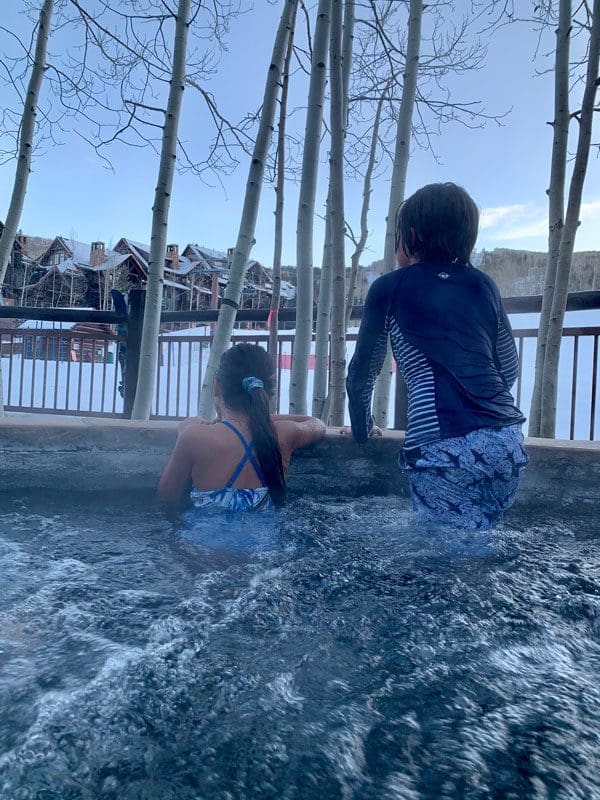 Two kids lean out of an outdoor hot tub to enjoy a wintery scene at The Ritz-Carlton, Bachelor Gulch.