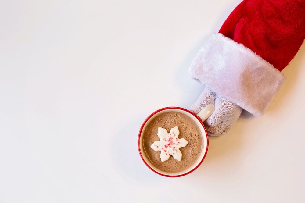 A Santa hand reaches out to hold a mug of hot cocoa with a giant, snowflake marshmallow.