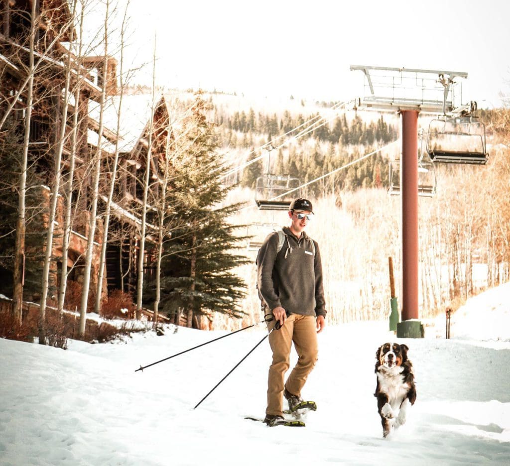 A man snowshoes in the snow with a large dog under several ski lifts. 