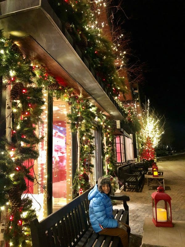 A young boy sits on a bench on a street in Vail, with storefronts well-lit and decorated for the holidays.