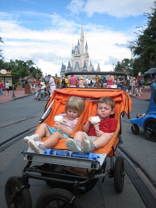 Two young kids enjoy ice cream in a double stroller, with Cinderella's Castle in the distance.