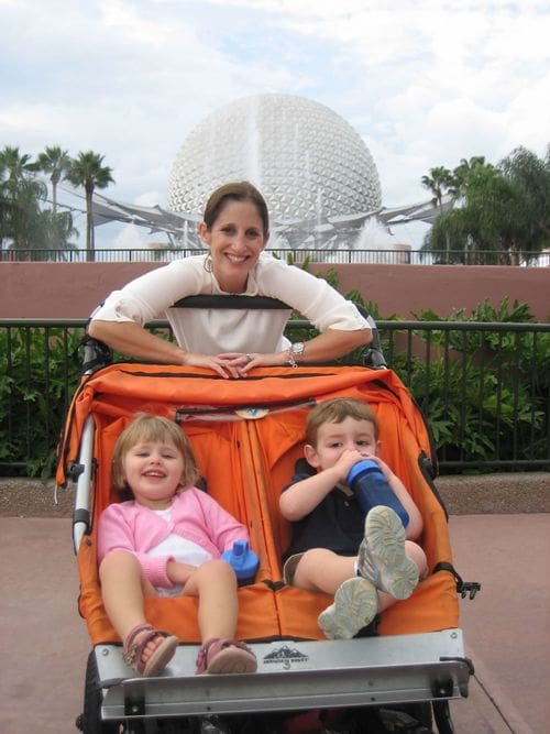 A mom stands behind a double stroller, holding her two young kids, with the iconic Epcot white ball behind them.