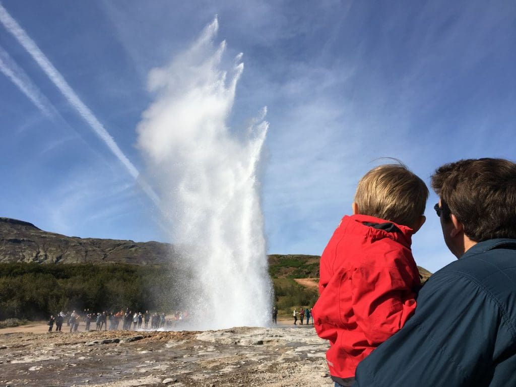 A dad holds his young son as they look at a geysir in Iceland.