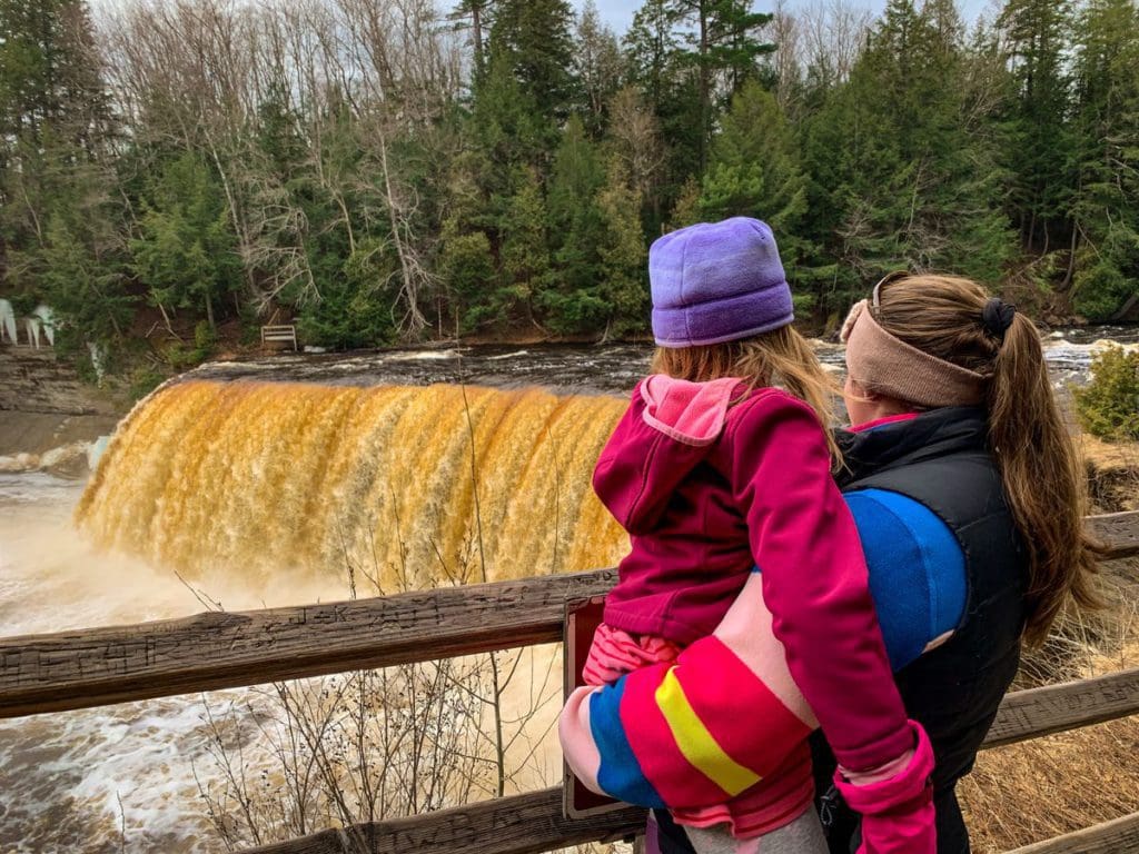 A mom holds her young daughter as they look at the Upper Falls at Tahquamenon State Park, one of the best Upper Peninsula Michigan hikes for families.