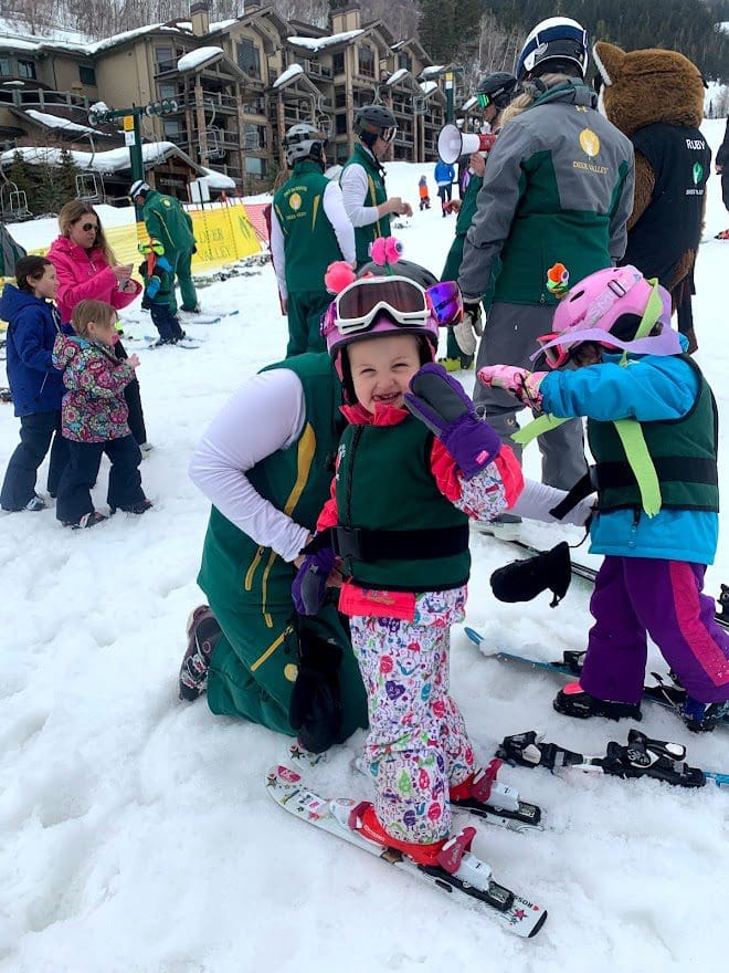 A toddler girl in skis and a full snowsuit prepares for ski lessons.