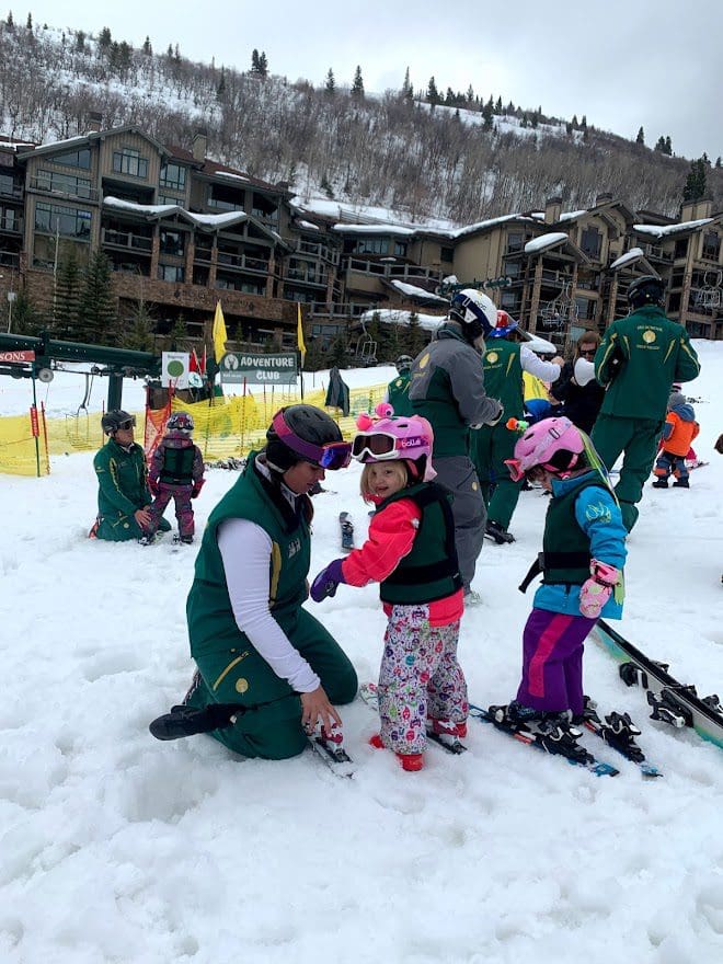 A young girl gets help putting on her ski gear from a ski instructor before a lesson.