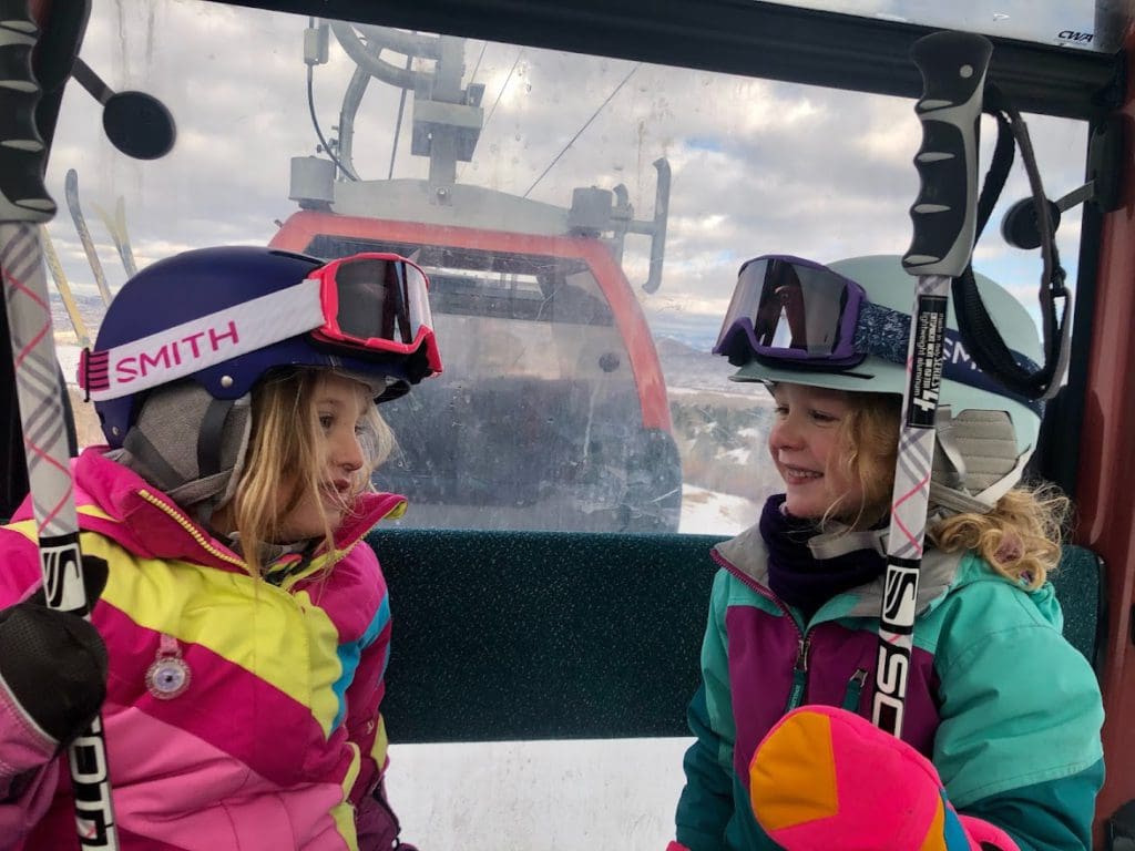 Two little girls in full ski gear and helmets smile at each other as they ride a gondola while visiting Deer Valley.