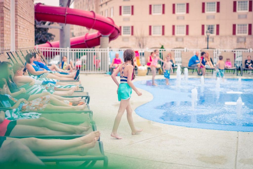 A young girl walks towards an outdoor pool, with a large slide in the background, at Hershey Lodge.