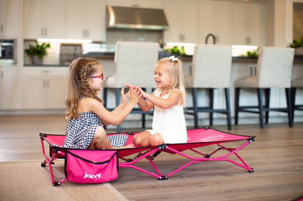 Two young kids sit facing each other playing a game on a Joovy Foocot Toddler Travel Cot.