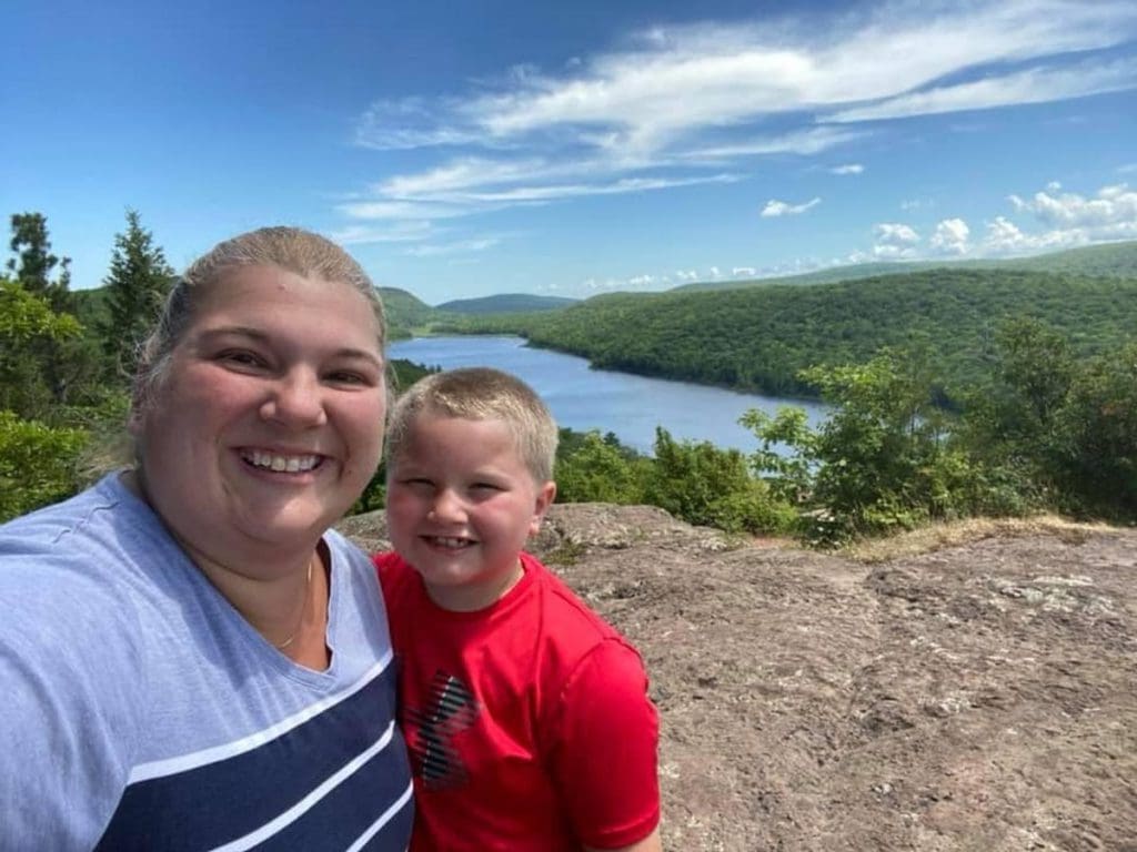 A mom and her son smile after hiking to the top of an overlook, with a view of the Lake of the Clouds behind them in the UP of Michigan, one of the best cool-weather destinations in the United States for families.
