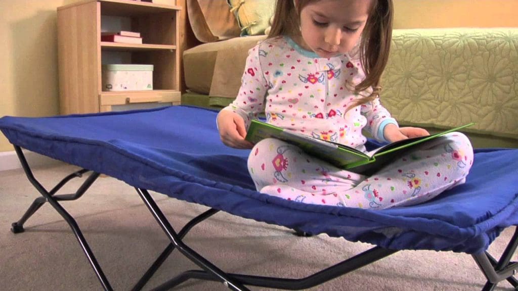 A young girl sits on a Regalo My Cot Portable Toddler Bed reading a book.