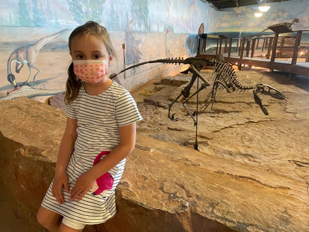 A young girl sits near an exhibit at the Dinosaur National Monument in Utah, with a small dinosaur skeleton behind her. 