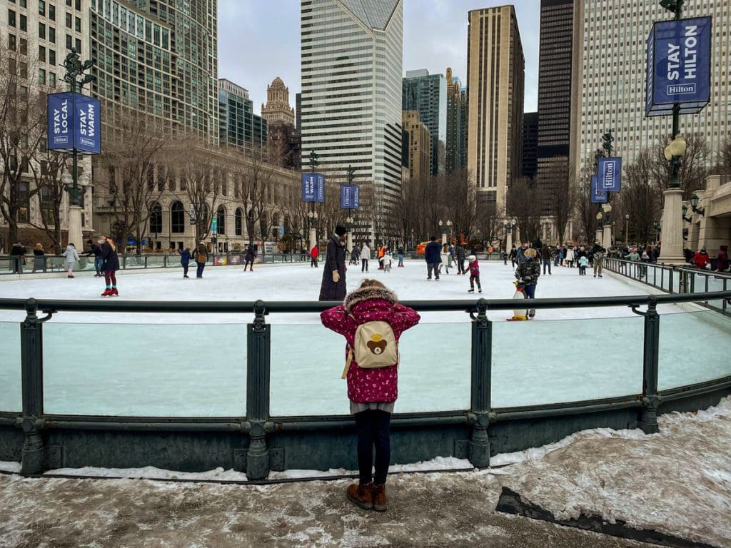 A young girl looks over a skating rink barrier at several skaters enjoying a winter day at Millennium Park.