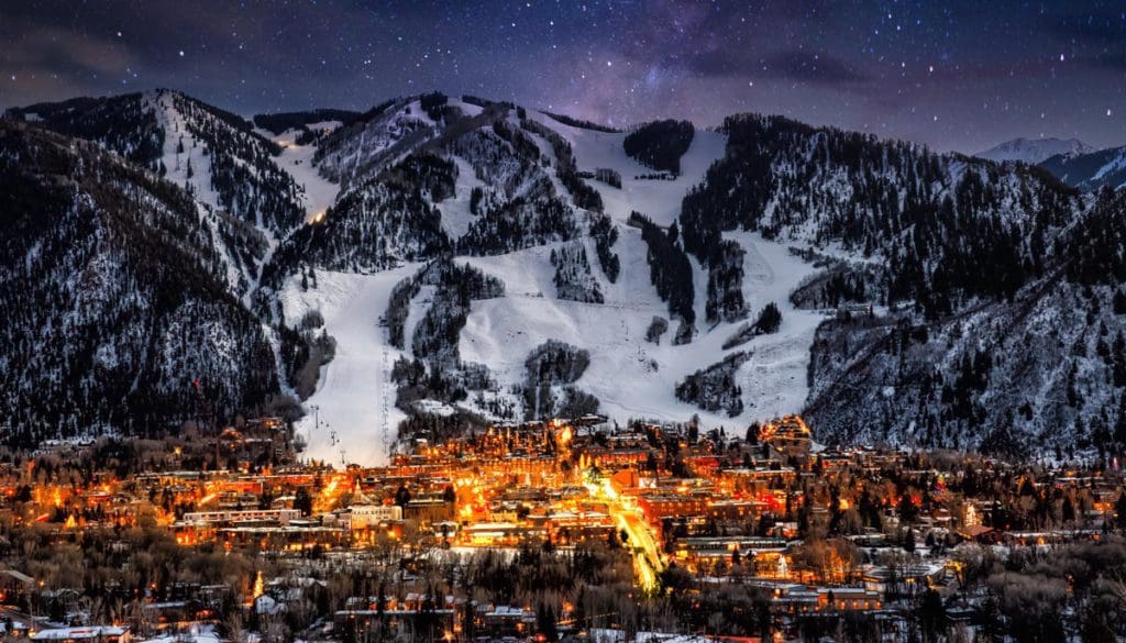 An aerial view of the downtown area of Aspen, lit up at night, with snowy mountain slopes in the distance.