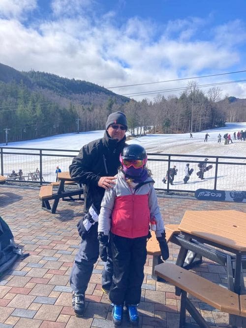 A dad and his young daughter, in full ski gear, stand at the patio of Whiteface Mountain Ski Resort.