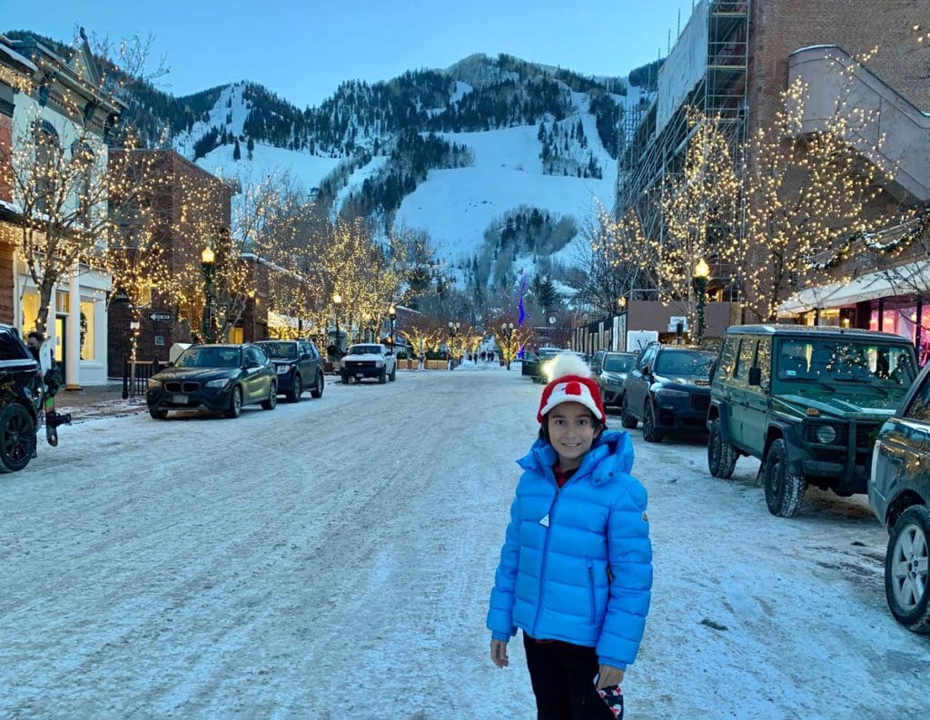 A young boy stands on a snowy street with a view of downtown Aspen behind him, and snowy mountains in the distance.