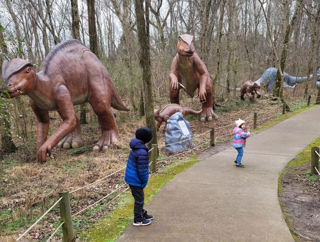 Two kids wander a path at Dinosaur World, while several Dinosaur replications stand on the other side of a rope fence.