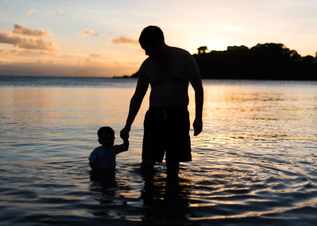 A dad and his young son walk in the water at sunset.