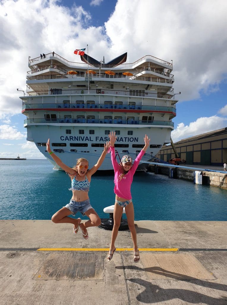 Two girls jump excitedly with a Carnival Cruise ship behind them.